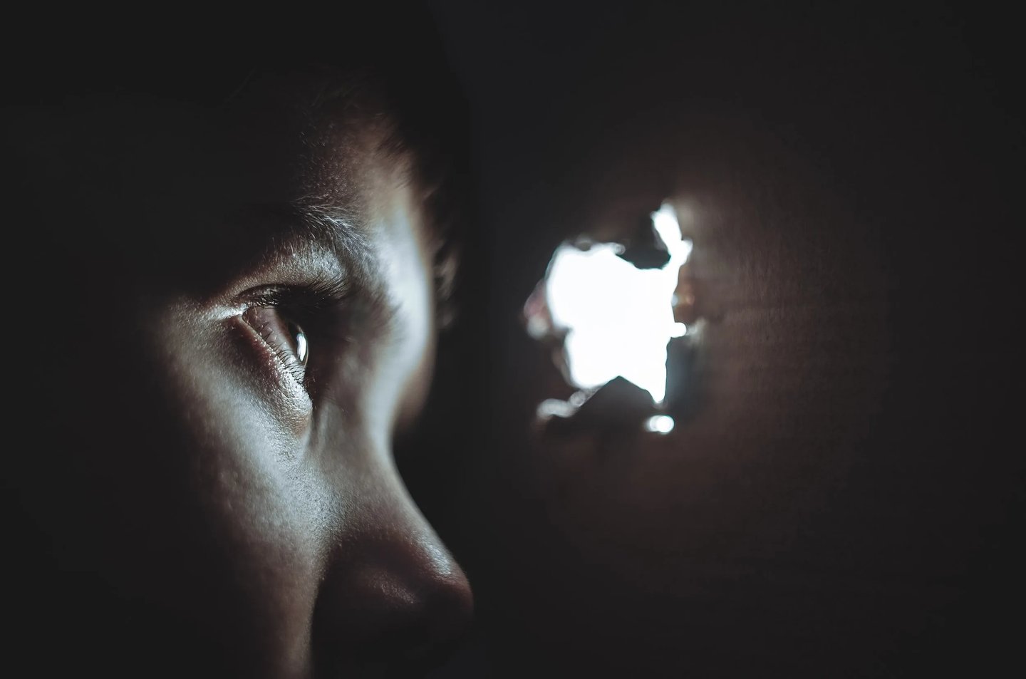 Close-up of a child's eye in darkness, peering toward light shining through a hole